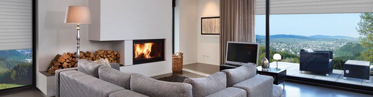 Modernes Wohnzimmer mit grauem Sofa, Kamin, Holzstapel, großem Fenster und Panoramablick auf Hügel und Stadt.